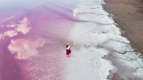 AERIAL, top view: joyful young woman in red skirt runs on water along white salty shore of amazing pink lake with clouds reflections. happy carefree female in vacation in wild nature. clip has noise