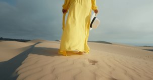 Close-up of woman in flattering yellow dress walking barefoot by wavy sand dunes in desert landscape. 4K Slow Motion golden sunset light highlighting model. Slim female legs with nature background