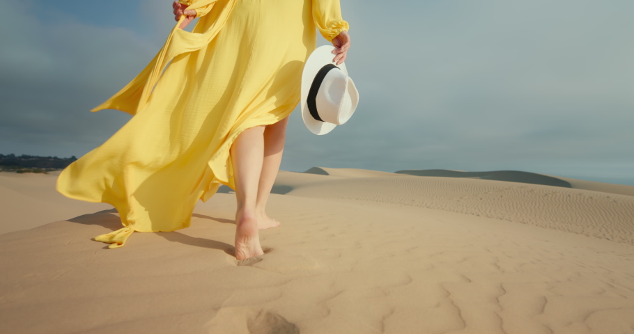 Close-up of woman in flattering yellow dress walking barefoot by wavy sand dunes in desert landscape. 4K Slow Motion golden sunset light highlighting model. Slim female legs with nature background