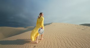 Cinematic slow motion of woman walking by sand dune. Barefoot female traveler in waving on the wind dress by the rippled sand desert surface with dark grey cloud on background. 4K Scenic nature 