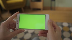 Young Woman Home Sitting on a Couch with Green Screen Smartphone in Horizontal Mode. Girl Using Touch Screen Mobile Phone. Girl Using Smartphone, Browsing Internet, Watching Video Content, Blogs. POV.