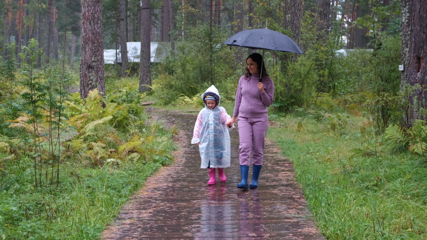 Little Girl in Raincoat with Mother Enjoying Their Walk on Wet Woden Path in Autumn Rainy Day. Slow Motion. Harmony with Nature and People concept