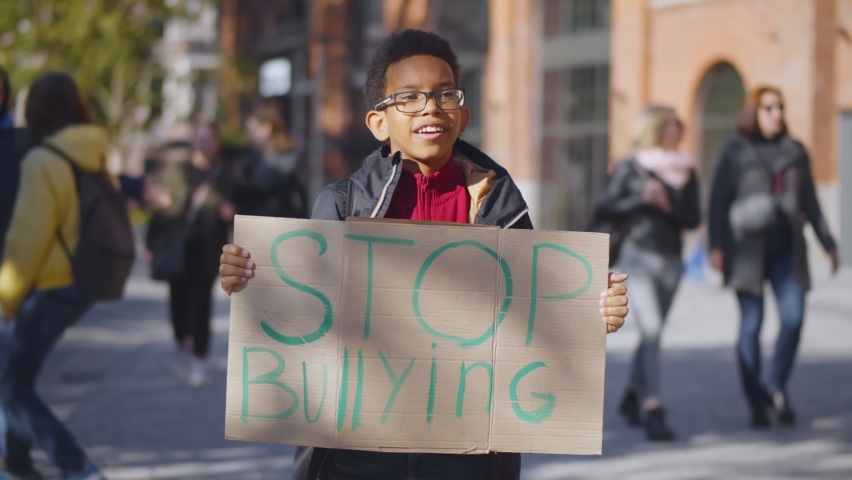 Portrait of african preteen boy in eyeglasses standing outdoors school building with stop bullying cardboard sign. Bully, aggressive behavior and offence concept Royalty-Free Stock Footage #1060523782