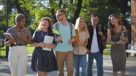 Multiethnic group of young cheerful students walking and talking outdoors. Diverse college friends with backpacks and books walking in campus park after classes