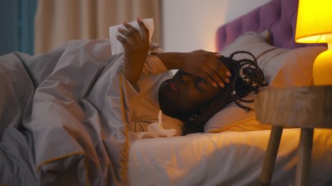 Sick Afro Woman Coughing in Tissue Lying In Bed Covered With Blanket At Home. Close up of young ill african female sneezing using paper handkerchief lying in bedroom