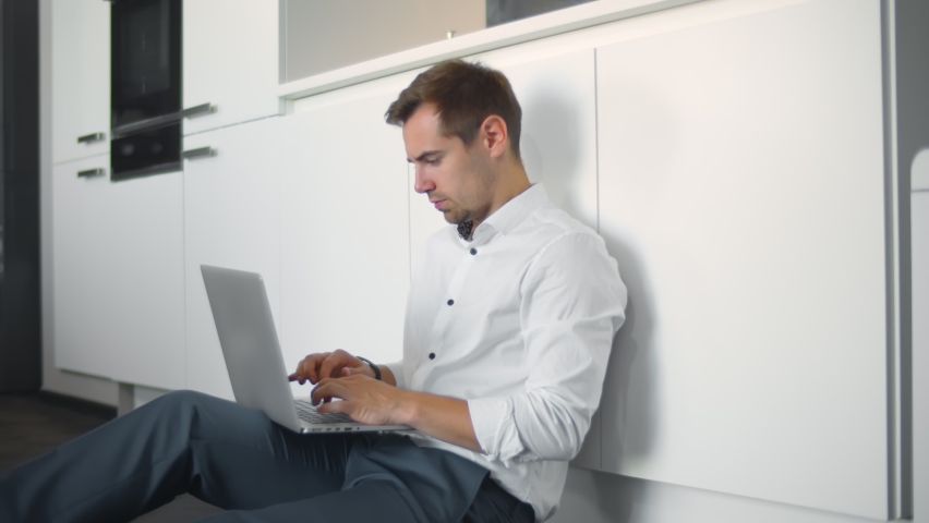 Young caucasian businessman working from home sitting on kitchen floor using laptop. Portrait of handsome man in formal wear relaxing on floor in modern home kitchen surfing internet on computer | Shutterstock HD Video #1060524286