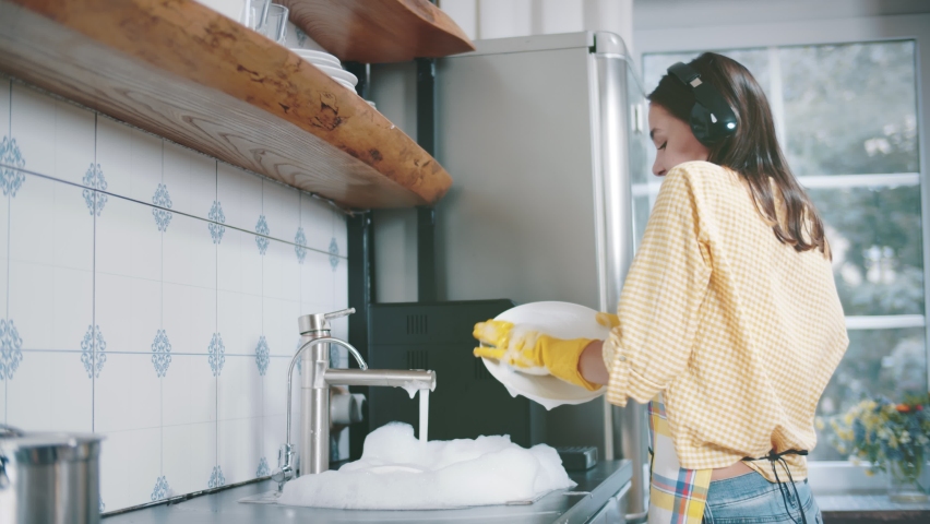 Side view of attractive young woman washing dishes listening to music in headphones in kitchen. Pretty cheerful housewife in rubber gloves wash plates enjoying music in earphones