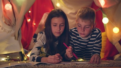 Happy preteen boy and girl drawing together while lying in play tent. Portrait of adorable brother and sister kids having pajama party and painting lying in wigwam with lights garland