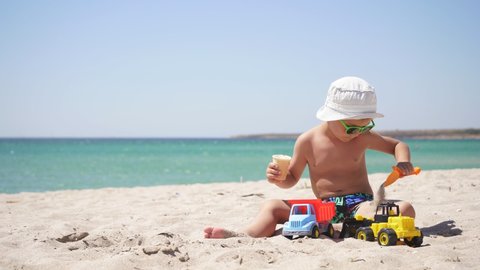 on a Sunny hot summer day, a boy plays with sand and toys, cars on the beach. child eats ice cream sitting on a sandy beach against the background of the azure sea.