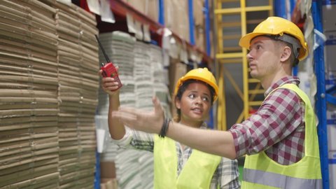 Young African female and Caucasian male warehouse workers in yellow reflective safety vest and yellow helmet are checking the goods with the other hand holding walkie-talkie in the warehouse.