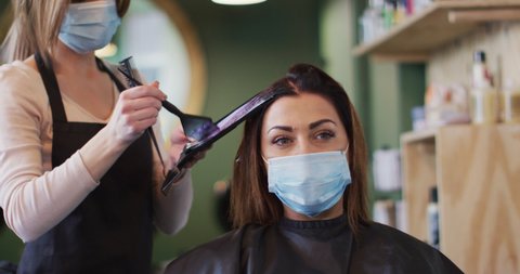 Caucasian female hairdresser working in hair salon wearing face mask, dying hair of Caucasian woman in face mask, slow motion. Health and hygiene in workplace during Coronavirus Covid 19 pandemic.
