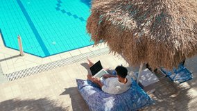 Man Greets and Talks Online by Internet Video Call Using Laptop in Resort Hotel Swimming Pool. Businessman Works on Vacation in Summer. Male Freelancer with PC Outdoors. 4K Handheld Wide Shot
