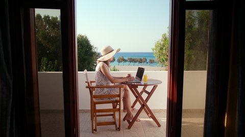 Young Woman in Dress and Hat Works on Laptop in Hotel Room Balcony with Sea View in Summer Sunny Day. Female Freelancer, Remote Worker or Businesswoman Uses Computer on Holiday. 4K Wide Handheld Shot