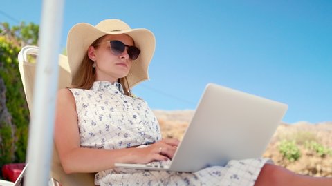 Beautiful Woman Works Using Laptop on Sea Beach Sunbed in Summer. Young Lady in Hat and Sunglasses on Vacation with PC. Female Freelancer or Remote Worker. 4K Handheld Medium Shot