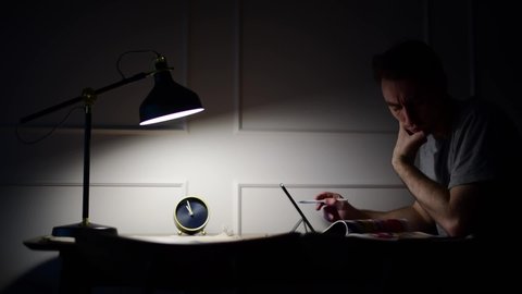 Serious concentrated young male student studying using tablet, making notes, busy man working late on pc at home office sitting in cozy workplace at night under lamplight.