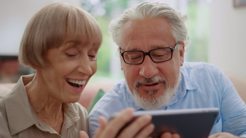 Smiling grandparents talking online on video chat with grandchildren. Senior people chatting on digital tablet at home. Mature couple using tablet. Happy woman sending air kisses at tablet camera
