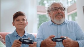 Focused grandfather playing video game with grandson at home. Mature man and boy using game joysticks. Senior man winning in computer game. Happy grandparent celebrating victory. Grandson losing game