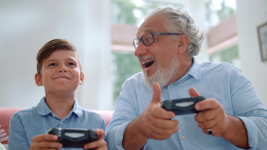 Focused grandfather playing video game with grandson at home. Mature man and boy using game joysticks. Senior man winning in computer game. Happy grandparent celebrating victory. Grandson losing game | Shutterstock HD Video #1060529650