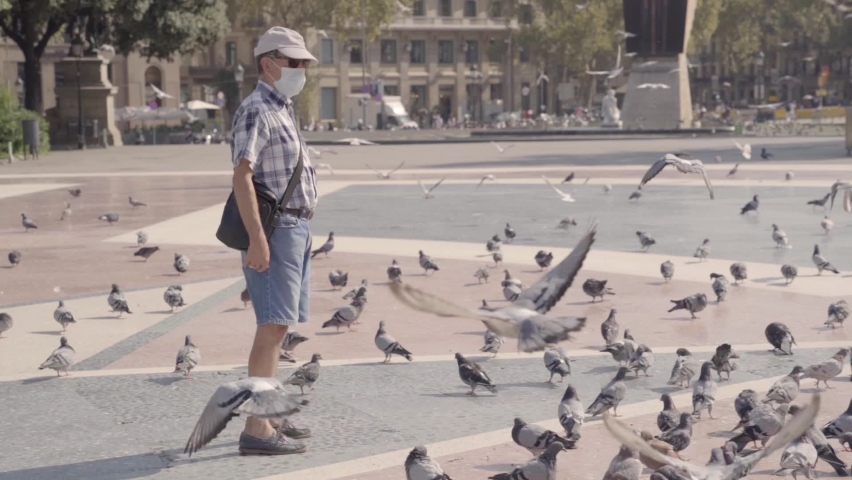 Old man with protective face mask on surrounded with pigeons in a public square