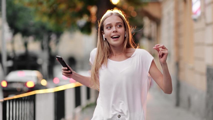 Young adult happy woman dancing in downtown very emotional. Slow motion of happy young woman in wireless headphones dancing singing outdoors in city street having fun alone