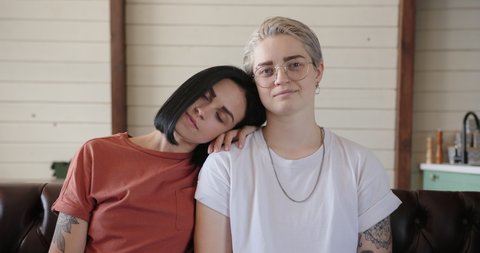 Serious lesbian couple with tattoos poses for camera sitting on leather sofa and hugging at home with white wall slow motion