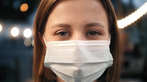 Close up portrait of caucasian young woman in medical mask standing at outdoor. Smiling person takes off mask. Healthcare. Safety measures concept. End of pandemic.