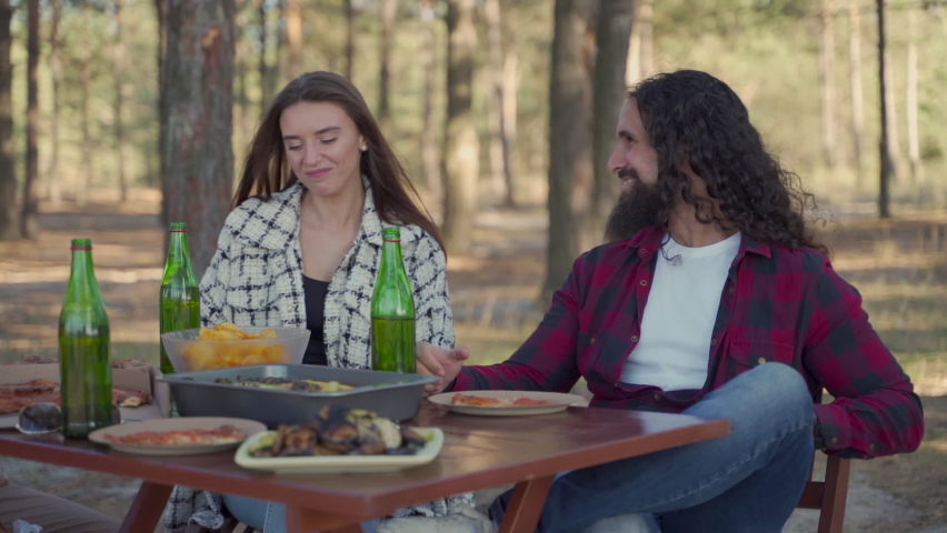 Young women and men sitting at a picnic table eating pizza and grilled food and drinking beer in nature