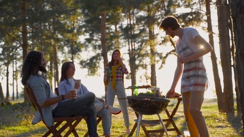 Young women and men cooking a barbecue and dancing in the countryside. People enjoying a picnic in nature.