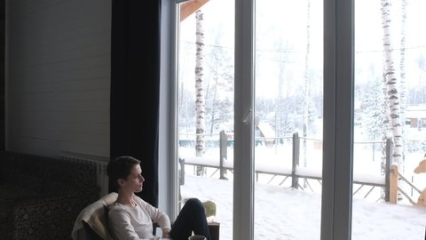 pretty girl with short hair sits on chair near large winter window barefoot and drinks coffee from Cup. enjoying hot drink. Winter landscape outside the window. Morning coffee in cozy home atmosphere