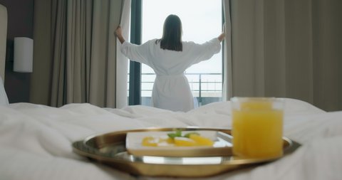 Young man with coffee cups comes to brunette woman in bathrobe opening window curtains in hotel room in morning backside view