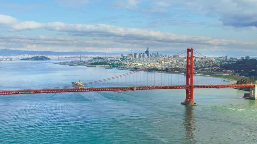 Aerial view of the magnificent Golden Gate Bridge with a cargo ship passing under it. This bridge connects the San Francisco peninsula to Marin County. US route 101 and SR 1 full of cars. Red 8K. Royalty-Free Stock Footage #1060541104