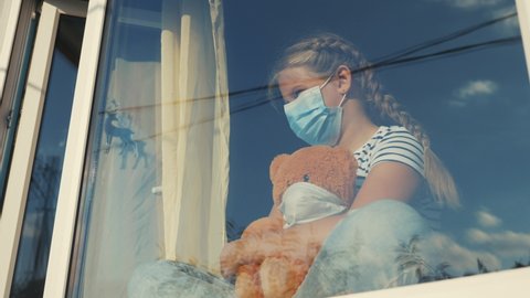 bored kid in medical mask in home quarantine coronavirus sitting by the window. child with a toy teddy bear in protective masks looking out the window. coronavirus epidemic prevention covid 19 concept