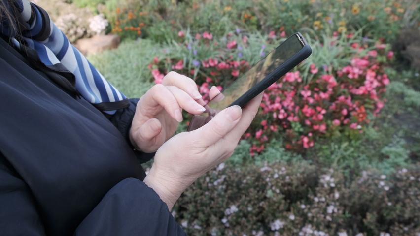 A woman in an autumn city park in a dark jacket holds a black smartphone in her hands and communicates on a social network, close-up, October cloudy day