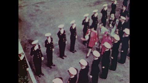 1940s: Flags wave near newly constructed ship. Military officers salute Admirals and their wives. Guillotine severs rope and ship glides down chute into harbor.