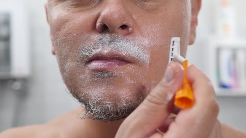 Male shaving with razor. Young man apply shaving foam on face in bathroom. Healthy skin, cares of body, white wall background.
