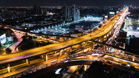 Time-lapse of car traffic transportation on highway, road intersection, sky train railway, with under construction site at night. Urban transport lifestyle, Asian city life. High angle cityscape view