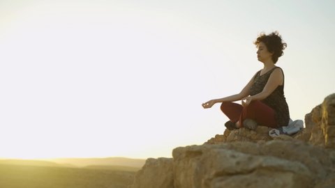 Young woman relaxing practicing meditation on a desert mountain top at sunset. Self examination, concentration and deep thought on a cliff with epic and beautiful colors. Sits quiet and peaceful.