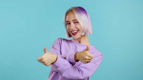 Cute woman with violet dyed hairstyle showing thumb up sign over blue background. Positive young girl smiles to camera. Winner. Success. Body language. 