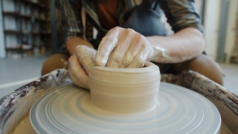 Close-up of man's hands designing and making bowl from clay in pottery workshop while throwing wheel is rotating. Creative people and hobby concept.