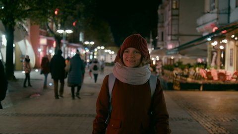 Portrait of Happy Young Woman Walking and Smiling to Camera Outside in Street in Late Evening or Night in Autumn or Winter. Tourist Lady in Red Jacket with Backpack. 4K Tracking Medium Shot