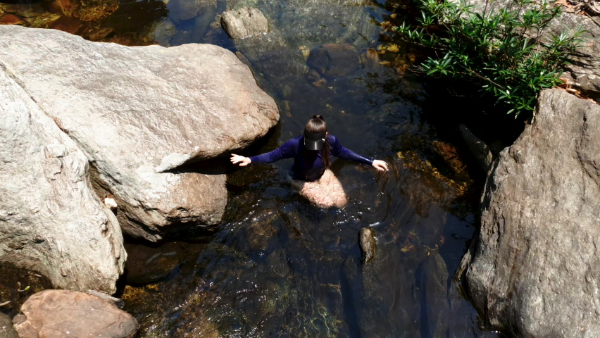 Young Latin woman in swimwear wading in rocky natural pool, medium down shot