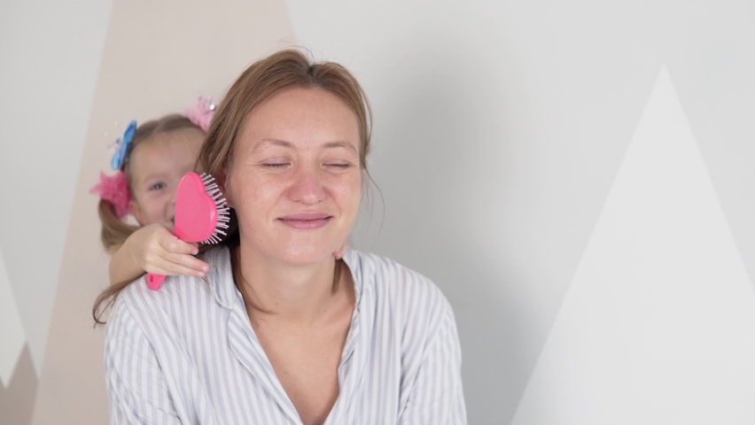 A little girl combs her mother's hair with a comb. Mom and daughter play  beauty salon at home.