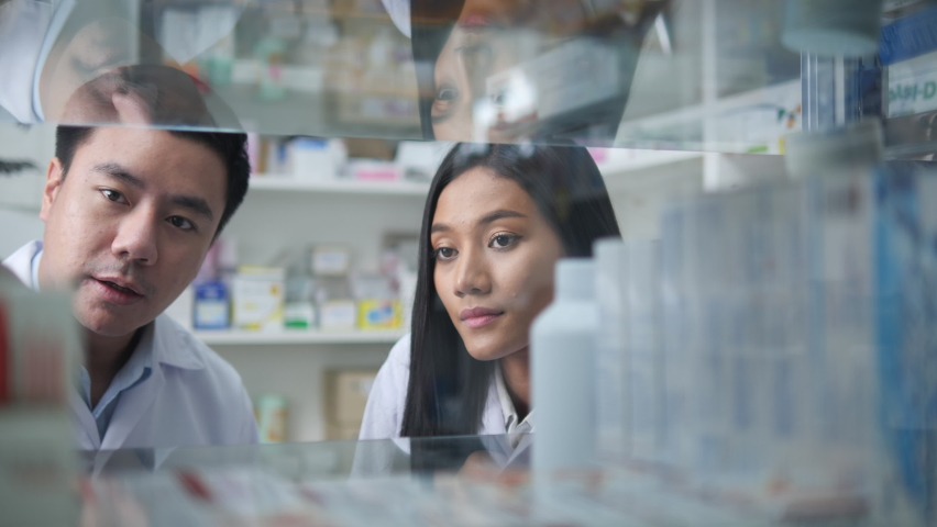 Asian pharmacists working and checking drug in pharmacy store at hospital. Asian doctor checking medicine cabinet and wearing white medical coat. Shelves pharmacy background in drugstore. Royalty-Free Stock Footage #1060574746