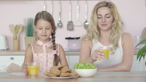 Happy Caucasian mother and daughter drinking healthful orange juice, talking, and smiling at camera. Portrait of positive young woman and little girl having healthy breakfast in the morning.