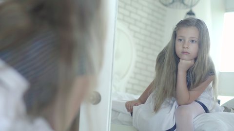 Sad little girl looking in mirror and sighing. Reflection of upset Caucasian brunette child with brown eyes sitting on bed at home and thinking. Unhappy kid alone indoors.
