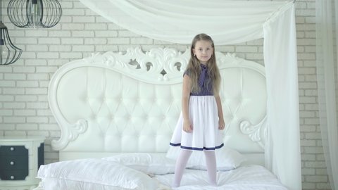 Wide shot portrait of cheerful brunette girl jumping on big white bed. Joyful Caucasian child having fun on weekends at home in bedroom. Charming kid enjoying morning indoors.