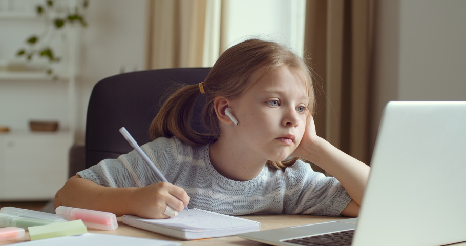 Portrait of preschool kid girl sitting at home, writing in notebook, studying doing homework, learning remote online in laptop app, child listening teacher with headphones, quarantine education study | Shutterstock HD Video #1060581388