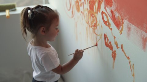 A little girl stands near the wall and draws with a paintbrush. portrait close-up
