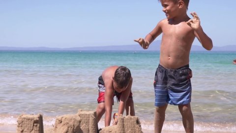 Two caucasian boys at beach of Stoupa, Peloponnese, Greece, one is building a sand castle and the other having fun dancing, slow motion medium shot