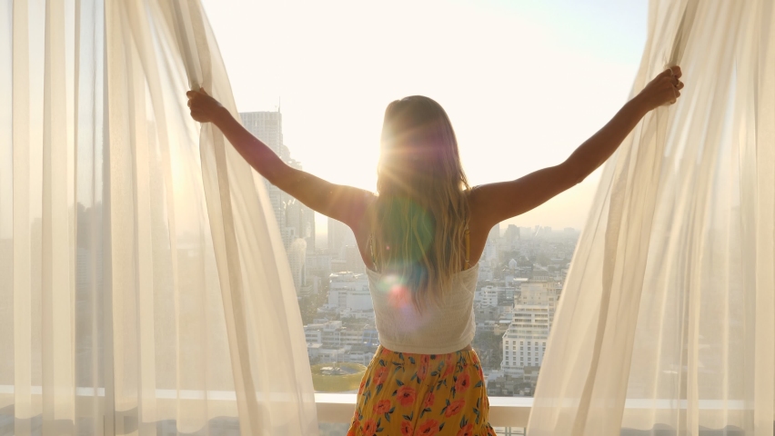Happy young woman opening curtain and looking through window at sun down. Smiling confident lady enjoying watching beautiful cityscape view from luxury hotel room  Royalty-Free Stock Footage #1060590442